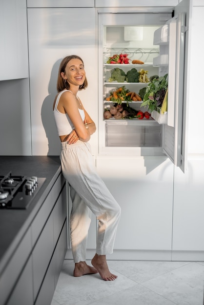 Mulher jovem em pé perto da geladeira cheia de legumes frescos na cozinha moderna. Conceito de alimentação vegana saudável