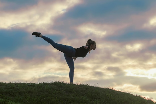 Mulher jovem em pé sobre uma perna e se exercitando na grama treino matinal, lindo nascer do sol