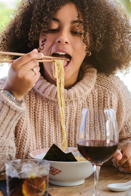 Mulher jovem em roupas casuais sentada em uma mesa de madeira com um copo de vinho e comendo macarrão com nori usando pauzinhos em um restaurante moderno