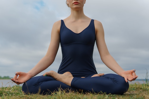 Mulher jovem em sportswear azul sentado em pose de ioga, céu nublado e lago no fundo.