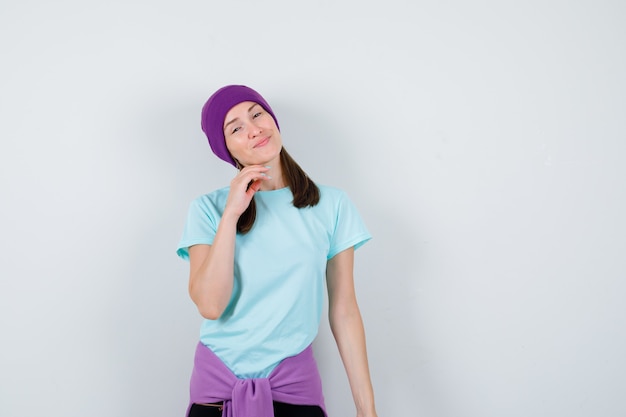 Mulher jovem em t-shirt azul, gorro roxo apoiando o queixo na mão, sorrindo e olhando alegre, vista frontal.
