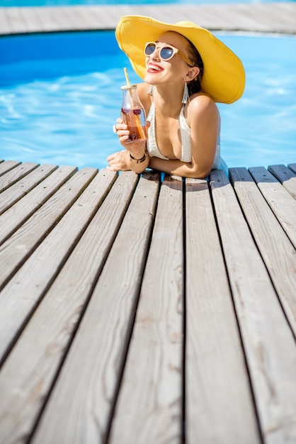 Mulher jovem em traje de banho com um grande chapéu de sol amarelo a relaxar com uma garrafa de uma bebida fresca, sentada à beira da piscina ao ar livre