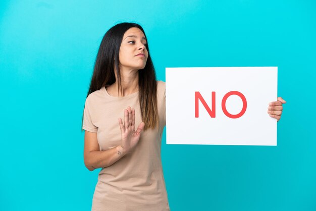 Mulher jovem em um fundo isolado segurando um cartaz com o texto NÃO e fazendo o sinal de pare