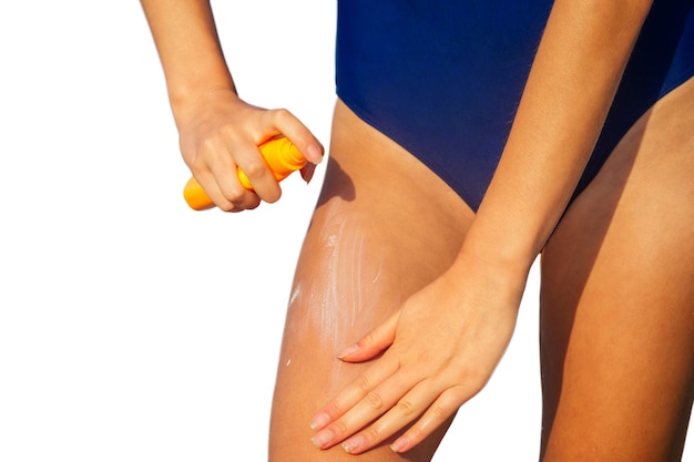 Mulher jovem em um maiô azul segurando um frasco de spray de embalagem de plástico laranja e colocando protetor solar no corpo. proteção ultravioleta do sol de verão FPS na praia isolada em fundo branco