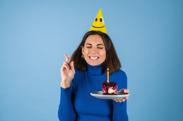 Mulher jovem em uma parede azul comemora aniversário, segura um pedaço de bolo, feliz, animada, faz um pedido