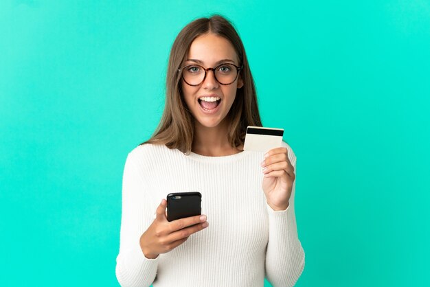 Mulher jovem em uma parede azul isolada, comprando com o celular e segurando um cartão de crédito com expressão de surpresa