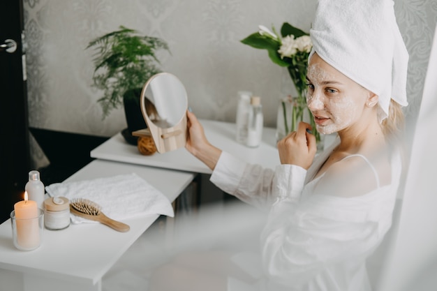 Mulher jovem em uma toalha branca relaxando no quarto e fazendo uma máscara facial de argila uma rotina de beleza, autocuidado