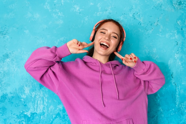 mulher jovem encantada com fones de ouvido, ouvindo música e dançando, isolada sobre uma parede azul dentro de casa