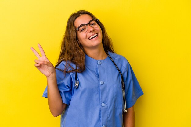 Mulher jovem enfermeira isolada em fundo amarelo alegre e despreocupada, mostrando um símbolo de paz com os dedos.