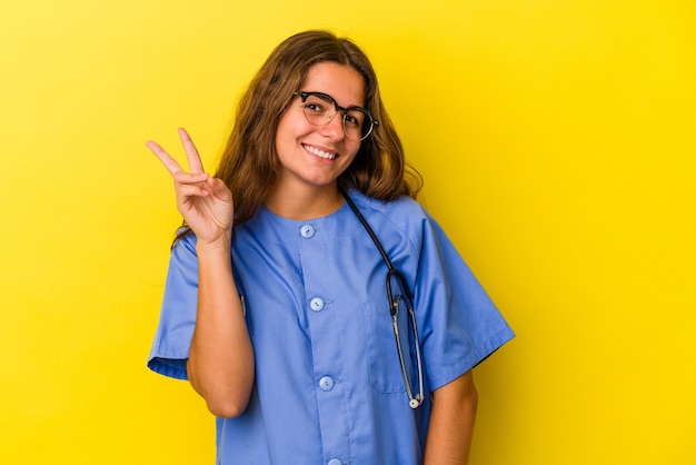 Mulher jovem enfermeira isolada em fundo amarelo, mostrando o número dois com os dedos.