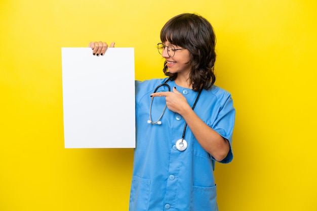 Mulher jovem enfermeira médica isolada em fundo amarelo segurando um cartaz vazio com expressão feliz e apontando-o