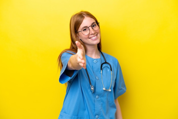 Mulher jovem enfermeira ruiva isolada em fundo amarelo, apertando as mãos para fechar um bom negócio