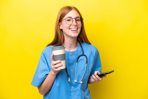 Mulher jovem enfermeira ruiva isolada em fundo amarelo segurando café para levar e um celular