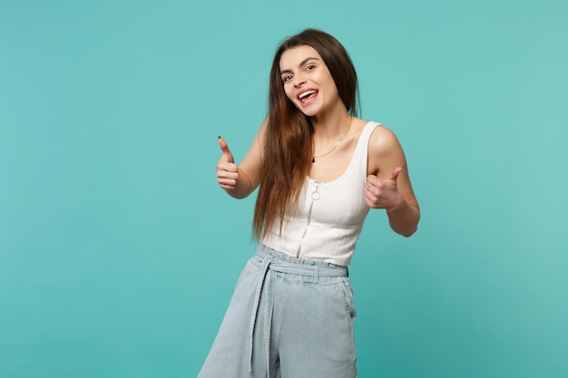 Mulher jovem engraçada alegre em roupas leves casuais, olhando a câmera, aparecendo os polegares isolados no fundo azul turquesa no estúdio. emoções sinceras de pessoas, conceito de estilo de vida. simule o espaço da cópia.