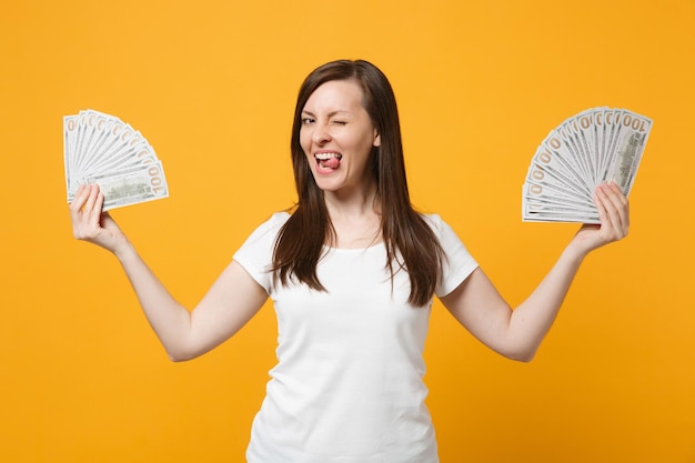 Mulher jovem engraçada em roupas casuais brancas mostrando a língua, segurando o ventilador de dinheiro em notas de dólar isoladas no fundo da parede laranja amarelo brilhante. Conceito de estilo de vida de pessoas. Mock up espaço de cópia.