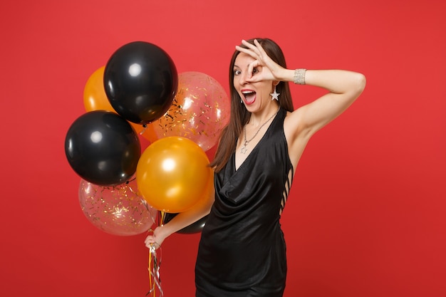 Mulher jovem engraçada em um vestido preto comemorando mostrando sinal de ok perto dos olhos segurando balões de ar isolados sobre fundo vermelho. Feliz dia internacional da mulher feliz ano novo, conceito de festa de feriado de maquete de aniversário.