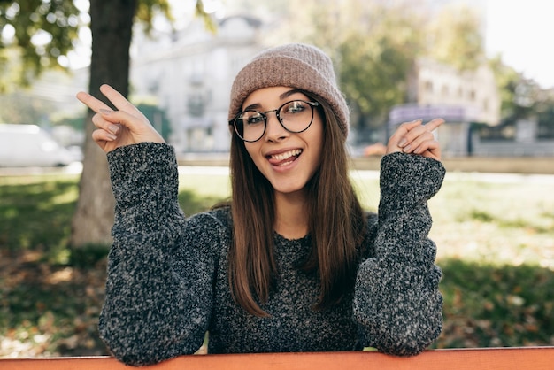 Mulher jovem engraçada sorrindo vestindo suéter e óculos transparentes na moda mostrando gesto de paz Linda jovem feliz relaxando lá fora na luz do sol no parque da cidade