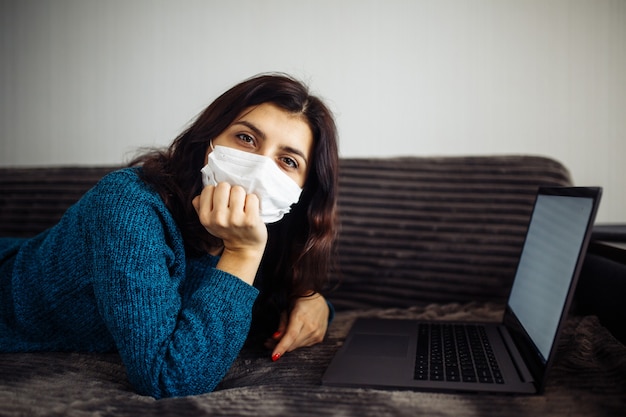 Mulher jovem entediada trabalhando em casa durante a quarentena devido à pandemia de coronavírus. Linda garota fica em casa usando máscara médica e digitando em um laptop. Conceito mundial de epidemia Covid-19.
