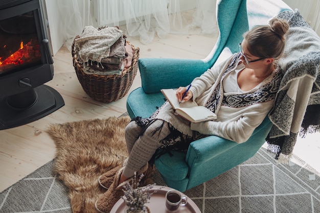 Mulher jovem escrevendo em um caderno, sentada em uma poltrona confortável perto da lareira