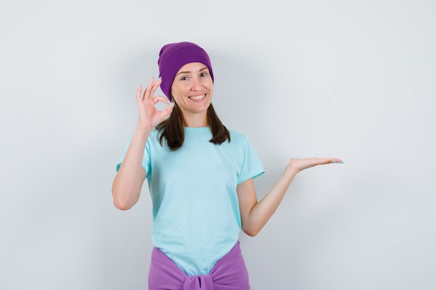 Mulher jovem espalhando a palma da mão de lado, mostrando sinal de ok em camiseta azul, gorro roxo e parecendo alegre. vista frontal.