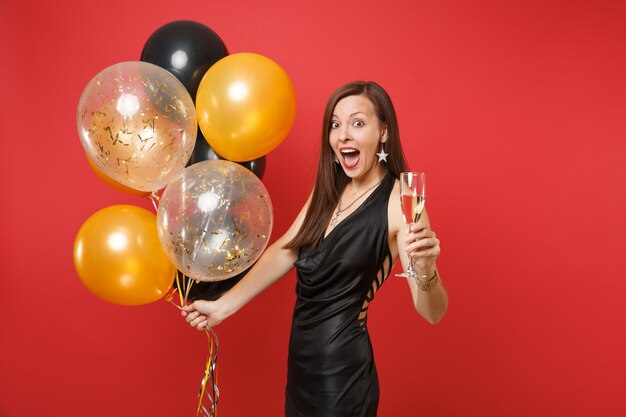 Mulher jovem espantada animada em vestido preto comemorando, segurando a taça de champanhe, balões de ar isolados sobre fundo vermelho. Dia de São Valentim, feliz ano novo, conceito de festa de feriado de maquete de aniversário.