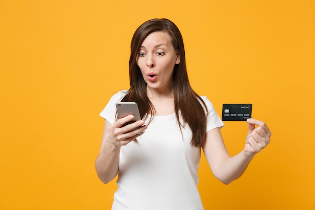 Mulher jovem espantada em roupas casuais brancas, mantendo a boca aberta, usando telefone celular, segurando o cartão bancário de crédito isolado em fundo laranja amarelo brilhante. conceito de estilo de vida de pessoas. mock up espaço de cópia.