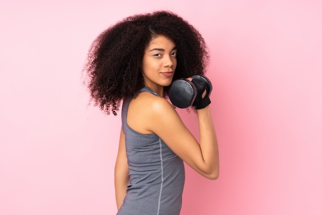 Mulher jovem esporte americano africano isolada na parede rosa fazendo levantamento de peso
