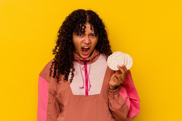 Mulher jovem esporte de raça mista segurando um bolo de arroz isolado no amarelo gritando muito zangado e agressivo.