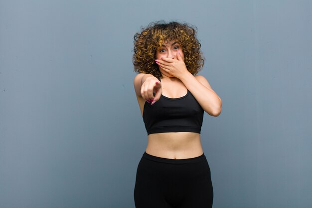 Foto mulher jovem esportes rindo de você, apontando para a câmera e tirando sarro ou zombando de você contra a parede cinza