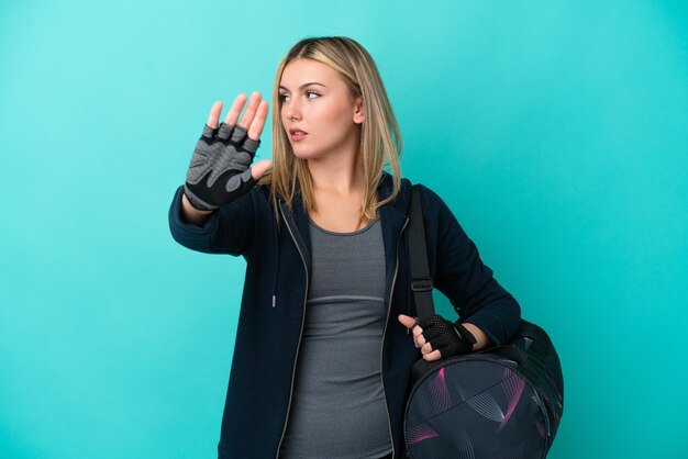 Mulher jovem esportiva com bolsa esportiva isolada em fundo azul fazendo gesto de parada e desapontada