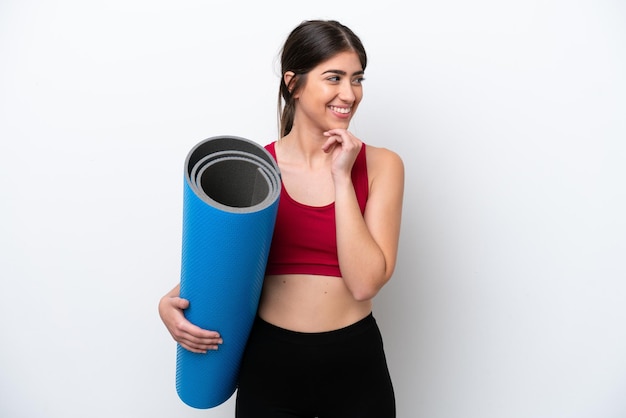 Mulher jovem esportiva indo para aulas de ioga enquanto segura um tapete isolado no fundo branco, olhando para o lado e sorrindo