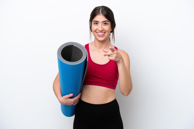 Mulher jovem esportiva indo para aulas de ioga enquanto segura um tapete isolado no fundo branco surpreso e apontando para a frente