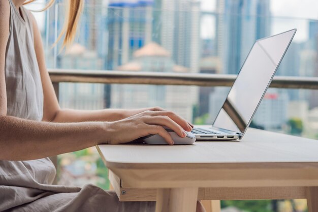 Mulher jovem está trabalhando em um laptop em sua varanda com vista para o trabalho remoto freelancer de arranha-céus