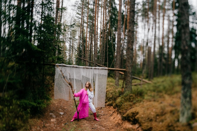 Mulher jovem estranha com capa de chuva rosa posando com um pedaço de pau na floresta