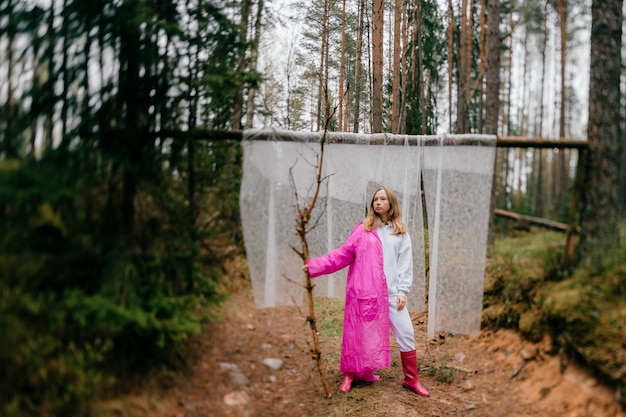 Mulher jovem estranha com máscara e capa de chuva rosa posando com um pau na floresta