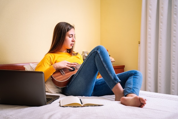 Mulher jovem estudando com ukulele em cima da cama com um laptop