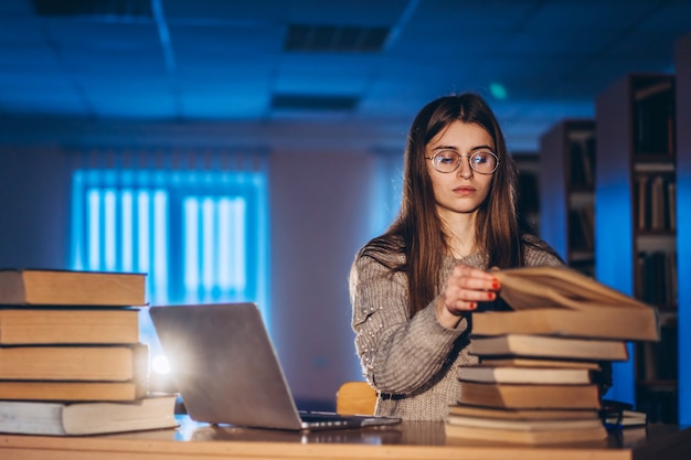 Mulher jovem estudante à noite se senta em uma mesa na biblioteca com uma pilha de livros e trabalha em um laptop. Preparando-se para o exame