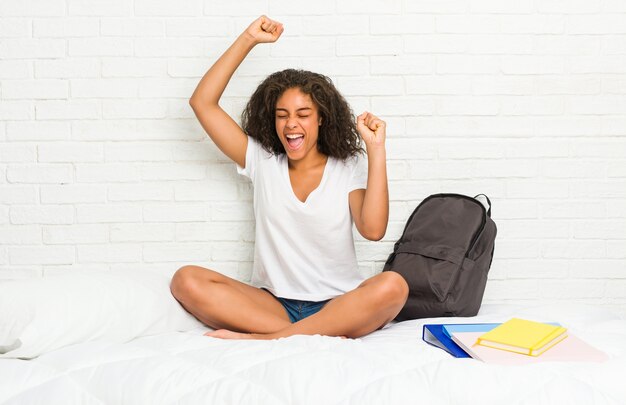 Mulher jovem estudante afro-americana na cama comemorando um dia especial, pula e levanta os braços com energia.