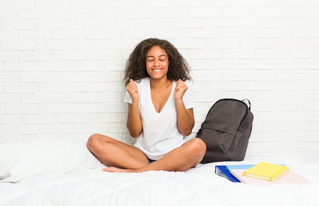 Mulher jovem estudante afro-americano na cama levantando o punho, sentindo-se feliz e bem sucedido. Conceito de vitória