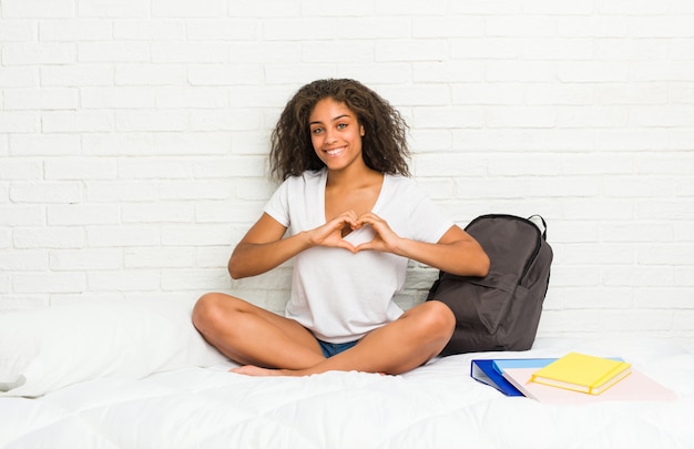 Mulher jovem estudante afro-americano na cama sorrindo e mostrando uma forma de coração com as mãos.