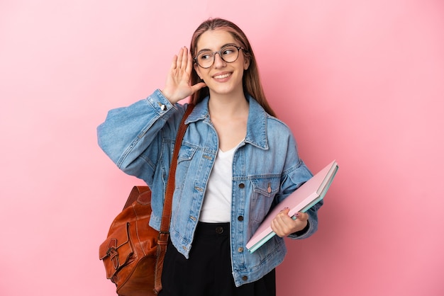 Mulher jovem estudante caucasiana isolada na parede rosa ouvindo algo colocando a mão na orelha
