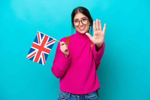 Mulher jovem estudante caucasiana segurando a bandeira inglesa isolada em fundo azul contando cinco com os dedos