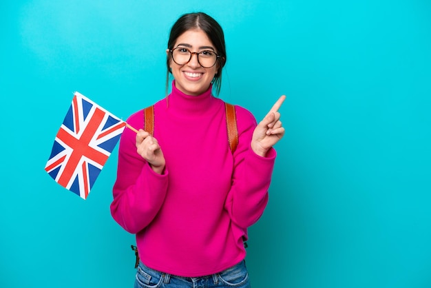 Mulher jovem estudante caucasiana segurando bandeira inglesa isolada em fundo azul, apontando para o lado para apresentar um produto