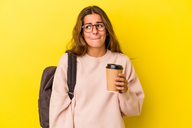 Mulher jovem estudante caucasiana segurando café isolado em um fundo amarelo confuso, sente-se em dúvida e inseguro.