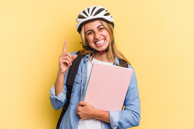 Mulher jovem estudante caucasiana usando um capacete de bicicleta isolado em fundo amarelo, sorrindo e apontando de lado, mostrando algo no espaço em branco.