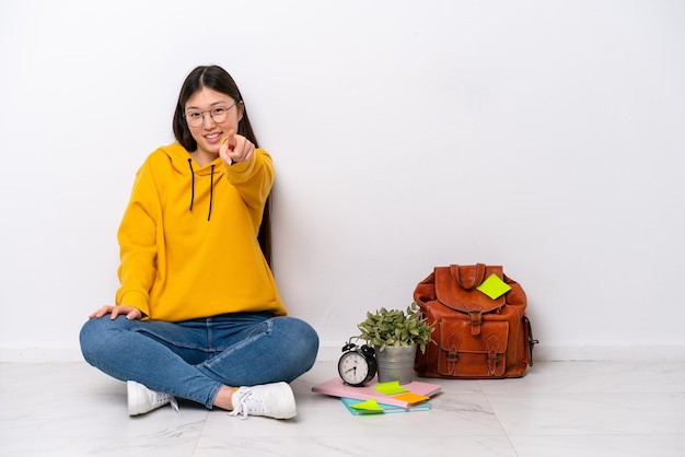 Mulher jovem estudante chinesa sentada no chão isolada na parede branca aponta o dedo para você com uma expressão confiante