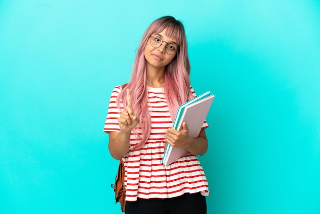 Mulher jovem estudante com cabelo rosa isolado em um fundo azul, mostrando e levantando um dedo