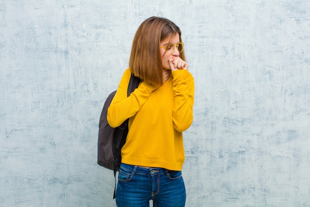 Mulher jovem estudante doente com sintomas de dor de garganta e gripe, tosse com a boca coberta contra a parede da parede do grunge