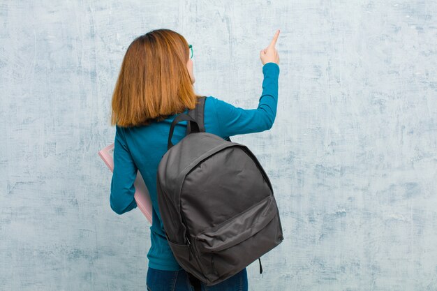 Mulher jovem estudante em pé e apontando para objetar no espaço da cópia, vista traseira contra o fundo da parede do grunge