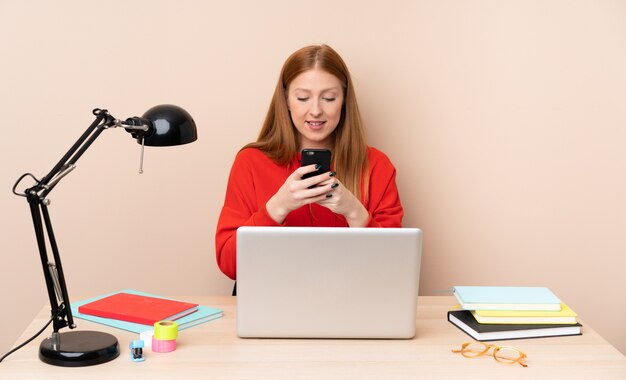 Mulher jovem estudante em um local de trabalho com um laptop, enviando uma mensagem com o celular